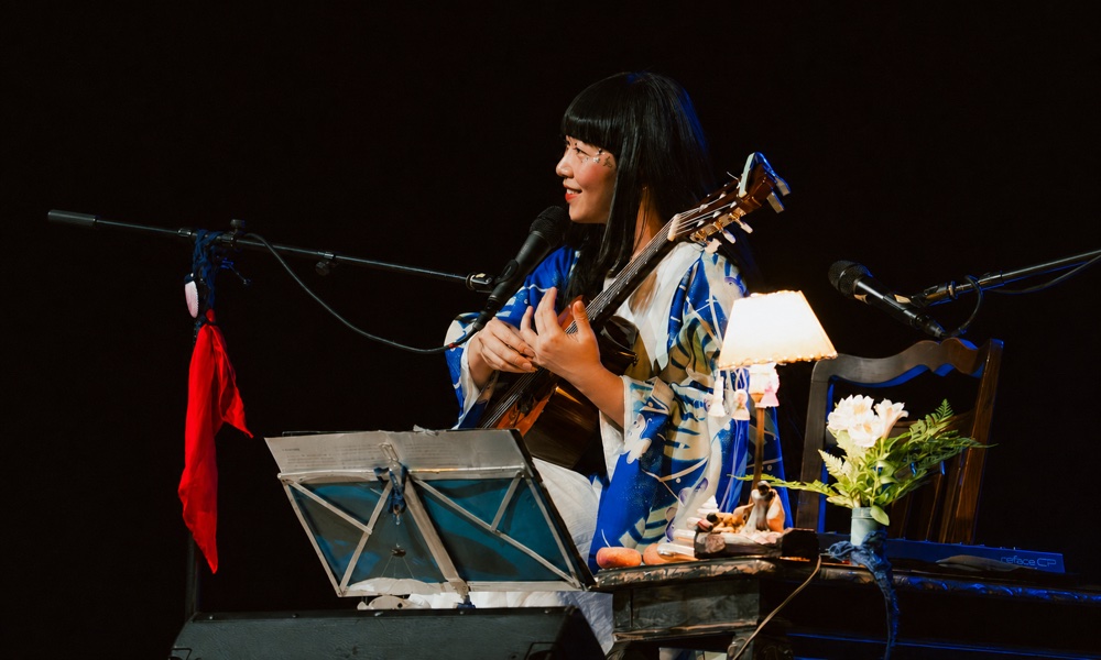 Ichiko Aoba tocando guitarra junto a una lámpara y flores sobre el escenario